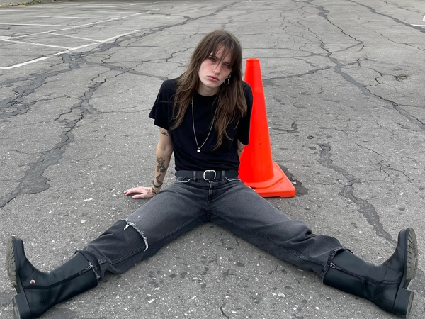 A young woman sits sprawled on rough, cracked asphalt in a deserted parking lot, framed straight-on in a casual iPhone mid-shot that feels perfectly timed yet unstaged. A bright neon-orange traffic cone rests provocatively centered between her wide-set legs, an ironic punctuation against the muted gray concrete. She wears a slightly oversized black crewneck tee that softly clings to her collarbones, paired with low-rise charcoal-gray jeans faded with subtle whiskering and a ripped knee, secured by a worn black belt with a matte silver buckle. Her matte black biker boots reach mid-calf with chunky soles and double straps, rugged and clearly well broken-in. Her long, tousled hair hangs in natural, greasy layers with parted bangs revealing part of her face, unstyled and raw.

Her skin carries the textured realism of iPhone photography—visible pores, faint redness, and natural imperfections with zero makeup or subtle traces melting away. She gazes through the lens with a dead-eyed, indifferent stare, chin tilted slightly upwards, exuding apathetic mid-2000s indie sleaze energy. Tattoos cover both hands, minimally detailed and partially visible. The flat, overcast daylight casts soft, even lighting, without filters or edits, emphasizing the rough textures of asphalt, faded fabric, and scuffed leather. The photo’s casual, no-nonsense composition and offbeat attitude create a genuine snapshot of gritty youth culture and defiant intimacy.
