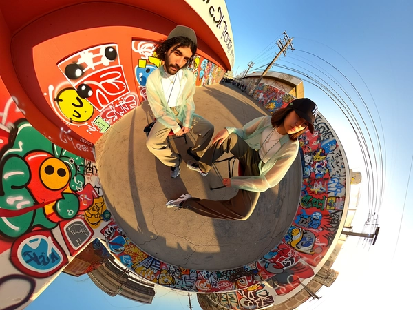 A spontaneous 360-degree candid image taken from a ground-level center point with an upward-facing spherical camera paired with an iPhone, capturing two models sitting casually on simple folding chairs in an empty lot covered with bold, colorful emoji-style graffiti. Both individuals wear effortlessly stylish sheer layered streetwear: lightweight translucent oversized jackets blending soft pastel hues over breathable tonal streetwear pieces, paired with relaxed-fit pants in muted earth tones, and chunky sneakers showing realistic subtle scuff marks. Accessories include minimalist silver chains and street-style caps worn loosely. Their hair is styled naturally with gentle imperfections, relaxed in candid poses—one leaning slightly forward, the other crossing legs, both minimally acknowledging the camera. The chaotic, vibrant graffiti walls envelop the scene, creating a swirling pop-art tunnel-like effect characteristic of the spherical fisheye perspective. Warm natural afternoon sunlight casts authentic dynamic shadows across the textured cracked concrete floor and highlights the intricate layering and fabric sheerness. Fabric textures reveal delicate mesh details, soft folds, and subtle translucency, while footwear shows nuanced wear. The immersive circular framing produces a dynamic, stylized tunnel wrapping the spontaneous urban scene, embodying the genuine casual yet fashion-forward essence typical of top-tier spherical 360-degree iPhone candid photography.