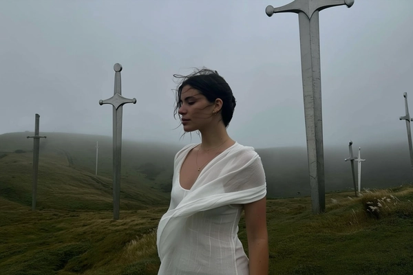 A spontanteously snapped, muted-toned iPhone portrait of a Greek woman standing pensively in a gently rolling hillside enveloped by soft mist and dotted with monumental, vertically embedded metallic swords. She wears a flowing, semi-sheer white summer dress subtly caught by a gentle breeze, the fabric and tall grasses swaying naturally around her. Her dark hair is lightly tousled by the humid air, partially damp and casually framing her quietly introspective face with authentic minimal makeup, revealing delicate skin textures and natural imperfections. The overcast grey sky diffuses light softly across the scene, casting muted shadows and reflecting faintly on the weathered surfaces of the monumental swords towering in surreal scale behind her. The casual, off-center framing includes a slight upward tilt emphasizing the subtle contrast between her calm stillness and the imposing, surreal sword structures, capturing an intimate, quietly melancholic moment within a mysterious, atmospheric landscape.