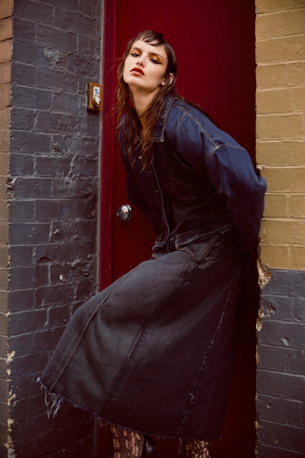 Dusky crimson light seeps into a grimy urban alley, wrapping a model poised against a timeworn brick wall painted in muted charcoal and ochre undertones. She wears a layered ensemble anchored by a jet-black denim skirt, its grainy surface roughened at the hem, paired with shredded stockings that reveal a tapestry of skin; her smudged eyeliner trails like whispered rebellion under cobalt shadows, while glossed lips catch flickers of mellow amber.

She leans with a quiet defiance, a moment caught between tension and ease, her gaze squinting through a measured haze at the camera, framed by loose strands that soften her angular jawline. The scrim of late afternoon light forms a chiaroscuro effect, its hardness veiled by a slight diffusion, coaxing every thread pull and abrasion in the denim, every pore and faint imperfection on her sculpted face into crystalline relief.

A narrow depth of field focuses tightly on the model’s upper torso, the brickwork dissolving softly behind in a tapestry of rough texture and urban decay, captured on a high-resolution digital sensor with a classic 85mm prime at wide aperture. The subtle digital noise renders the shadows with an analogue whisper, invoking the spirit of an archive rediscovered just in time.

Layers of warm umber and charcoal hues merge in the grading, lending the frame a timeless grit yet poised as a quiet manifesto of street-chic elegance—high-fashion editorial, hyper-real texture fidelity