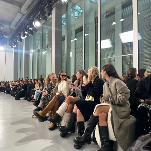 Four fashion girls seated front row, draped in oversized, textured outerwear paired with chunky designer boots, their expressions animated in mid-conversation as they lean into each other before the runway begins. The minimalist venue frames them with towering glass walls reflecting soft daylight, while scattered industrial beams and sleek concrete floors add understated edge. Natural light floods the space, casting soft shadows and subtle lens flares that mingle with slight digital noise from the iPhone sensor. The tactile contrast between knit sheens, leather scuffs, and cozy fabrics pops clearly, as a faint reflection of an LED panel flickers on a nearby window. Shot from a low-angle beside the runway aisle, the frame captures a candid intimacy, guests' murmurs blending into an ambient hum with a slight horizon tilt, edges cropped tight for editorial energy—contemporary runway snapshot, shot on iPhone