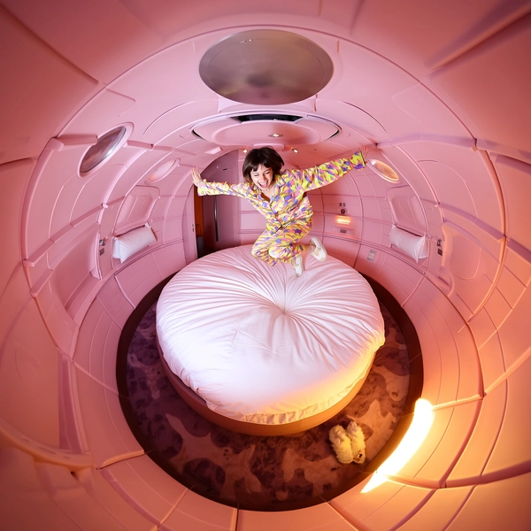 A spontaneous stylish 360-degree candid image inside a compact and whimsically designed pink capsule hotel room, the walls and ceiling swirling in a vivid fisheye tunnel-like distortion. At the center, a single person energetically jumps on a large round bed, dressed in a fluorescent pajama set with bold neon patterns and plush, furry slippers showing subtle wear. Their joyful, carefree expression and fluid mid-air posture convey genuine motion and relaxed spontaneity. Soft artificial lighting typical of cozy hotel interiors casts gentle shadows and reflects off smooth, textured bedding and sleek capsule surfaces. Detailed fabric textures highlight the silky pajama material's subtle sheen and natural folds, while the slippers' fuzzy texture and lightly scuffed soles add realism. The spherical, upward-facing ground-level camera viewpoint creates an immersive circular composition, emphasizing the playful circular bed and enclosing swirling pink capsule surroundings with authentic tactile and visual nuances. This image perfectly captures the distinctive qualities of casual, stylish, spontaneous spherical 360-degree photography achieved through a portable 360-degree camera paired with an iPhone, blending immersive tunnel-like distortions, realistic lighting, and fashionable casual energy.