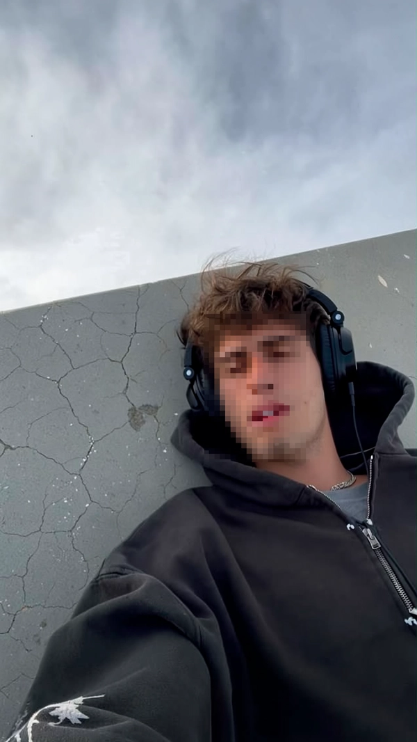 Casual candid image of a young man lying on cracked concrete outdoors, dressed in an oversized black hoodie with cigarette burn marks visible on the sleeve. He wears over-ear headphones and his face is deliberately pixelated to preserve anonymity and enhance mystery. The grey sky above softly diffuses natural daylight, casting realistic shadows and subtle highlights across his clothing textures and hair strands. The spontaneous framing captures a slightly tilted, close-up perspective with casual imperfections, reflecting an authentic, unscripted iPhone snapshot.