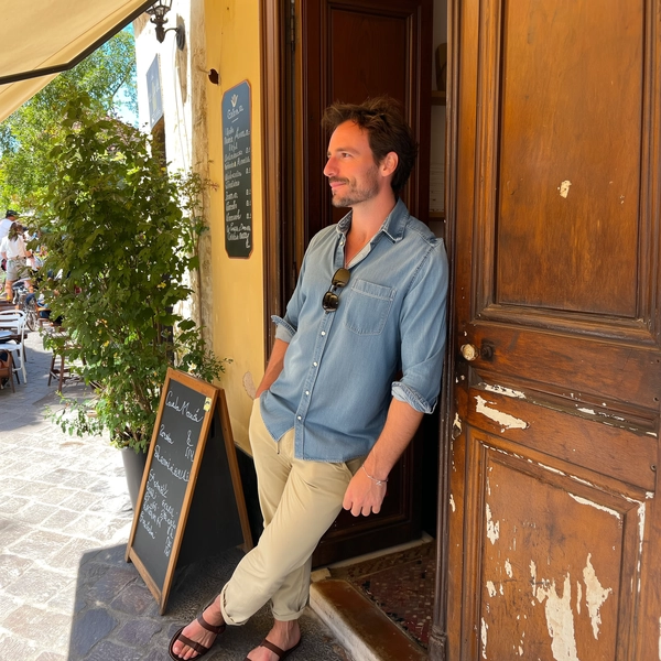 A candid photo of a Danish man casually leaning against the doorway of a rustic café in Modena, Italy. He wears a light denim shirt with the sleeves slightly rolled up and beige chinos, paired with simple leather sandals. His hair is short and slightly tousled, and he wears minimal accessories, including a delicate leather bracelet and classic aviator sunglasses resting on his collar. His expression is relaxed and thoughtful, gazing slightly off-camera as if enjoying a quiet moment. The café doorway features weathered wood with peeling paint and a small handwritten menu board visible beside him. Sunlight filters softly through nearby greenery, casting warm, natural daylight that highlights the textures of his fabric, textured stone pavement, and aged café walls. The composition is slightly tilted and framed spontaneously, capturing the easygoing elegance and genuine intimacy typical of casual iPhone snapshots in a lively Italian provincial town.