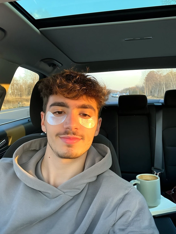 A young man sits comfortably reclining in the front seat of a car, hydrogel under-eye patches distinctly visible beneath his eyes. His hair is casually tousled, and he wears a soft, relaxed hoodie. Warm afternoon light streams through the windshield, softly illuminating the smooth texture of the patches and his natural skin tone. Nearby on the passenger seat, a ceramic travel mug with coffee rests casually beside an open notebook, adding intimate, personal detail. The selfie is taken from a natural front-facing angle typical of a handheld iPhone, clearly framing his face and shoulders in an unposed, genuine moment of self-care and calm—skincare selfie, shot on iPhone