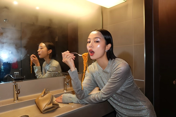 In the dimly lit confines of a sleek club bathroom, a young woman with impeccable, model-like poise sits perched beside a gleaming sink. Her shimmering silver glitter top catches the soft neon glow, casting delicate sparks across her smooth, meticulously styled hair that frames a serene, expression-neutral face. A subtle touch of highlighter accents her temples, and her lips hold a sheer gloss sheen that reflects the muted ambient light.

With measured grace, she lifts a fork twirling glossy noodles coated in a rich, spicy sauce, the strands glistening under the overhead spot. Her mouth parts just enough to accommodate the bite, eyes half-lidded, relinquishing no overt smile though a soft, gentle energy seems to ripple beneath her calm demeanor. The cool porcelain contrasts with the textured leather clutch casually draped from the edge of the sink nearby.

A casual iPhone shot captured from a slight knee-level tilt reveals the intimate scene: muted tiles fade into shadow behind her, while the faint haze of fogged mirrors glimmers in the warm neon halo. Tiny droplets linger on the fork’s gleaming prongs, and the temperature from the club’s late-night ambiance wraps the moment in tactile warmth.

This composition blends a candid yet editorial edge, knitting together the sparkle of nightlife with refined stillness. The understated harmony of polished textures and restrained movement culminates in a snapshot impeccably mirroring a spontaneous iPhone capture.