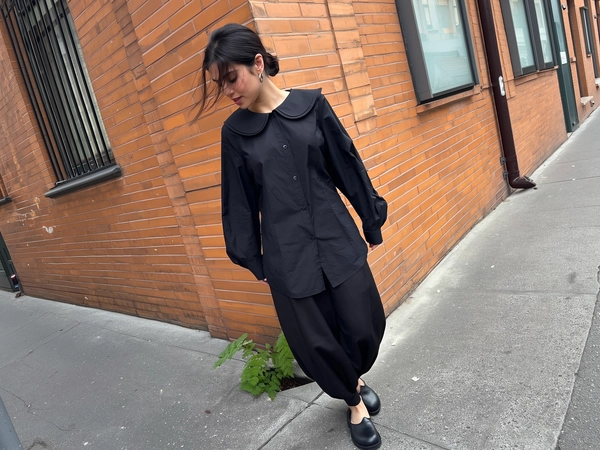 A spontaneous candid iPhone photo showing a young woman turning the corner of a warm-toned brick building on an urban sidewalk with a small green plant emerging from a crack nearby. She wears a thoughtfully Japandi-styled black button-up blouse featuring an exaggerated collar and oversized sleeves, neatly tucked into sculptural balloon trousers that taper gracefully at the ankle. Her rounded, thick-soled shoes add subtle grounded contrast. Soft natural daylight gently illuminates the scene, revealing tactile fabric grains, natural skin textures with subtle imperfections, and fine hair strands loosely framing her face. The casual framing is slightly tilted, creating an intuitively candid and relaxed perspective reminiscent of genuine street photography in a minimalist urban setting with textured concrete, aged brick, and delicate greenery accents.