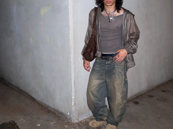A young man stands casually in a grimy concrete corner reminiscent of a back alley in an underground parking garage or half-abandoned gallery, framed in a slightly off-center mid-shot with an iPhone's subtle grain and soft focus. The cracked, gray splotched wall behind him bears faded graffiti and patches of water stains, while the stained floor shows tire marks and uneven wear. He wears an ultra-thin, crumpled taupe jacket with a dull sheen, layered over a faded charcoal-gray tight tee stretched at the collar. His oversized vintage bootcut jeans feature intricate embroidered dragons on one leg, fraying along the hem, dragging near dirty beige skate shoes, worn and blurred at the edges. A tangled cluster of gunmetal chains with random pendants hang loosely around his neck, visible beneath a mess of dark curls peeking out from a lived-in hoodie or jacket collar. A slouchy brown leather bag rests over one shoulder, completing the understated, indie sleaze look.

His pose is relaxed and unposed—one hand tucked in a pocket, the other hanging loosely as if caught mid-decision to light a cigarette, with his face cropped just above the lips, maintaining an air of mystery. Skin shows natural texture and subtle shadows cast unevenly by dim, ambient 3 a.m. lighting, reminiscent of an unfiltered iPhone 7 capture, with slight motion blur and imperfect framing that emphasize the raw immediacy. Tattoos, faint and line-based, peek from both hands, while the worn fabrics and frayed details evoke a lived-in authenticity. The overall feel is that of accidental cool—grunge softened with hints of Y2K vanity, captured with genuine imperfection and intimacy.