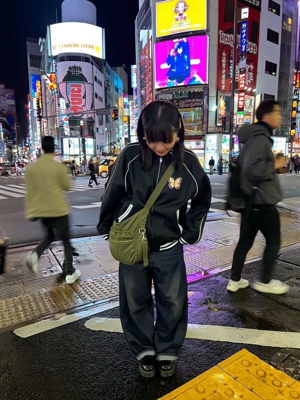 In Akihabara’s neon-splashed nightscape, a young woman leans slightly forward near a busy crosswalk, her oversized black track jacket detailed with faint stripes and a delicate butterfly patch catching cool street hues. Loose dark denim pools over chunky black loafers grounded on reflective wet asphalt, while an olive-green messenger bag slings casually across her torso, blending utilitarian charm with Tokyo’s gritty city pulse. Around her, radiating anime billboards and buzzing LED signboards flood the scene with fractured color, their vibrant glow casting soft halos over her lowered gaze. The iPhone’s lens hovers at hip-level, capturing faint lens flare and off-center framing as pedestrian silhouettes blur in motion behind—a candid urban moment swelling with the city’s restless hum. Textured jacket fabrics and slightly scuffed leather contrast with smooth neon brilliance, the whole scene tinged with grainy sensor warmth that holds quiet energy in Tokyo’s night sweep. —Tokyo street snap, shot on iPhone
