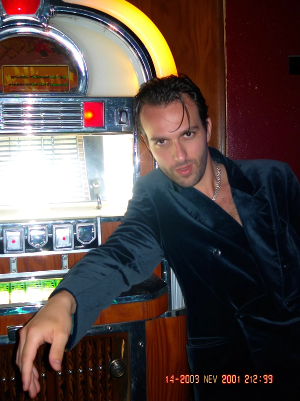 The flash bursts from the built-in lamp, throwing harsh white light across the slicked-back hair and velvet blazer of the man leaning casually against the retro jukebox. Chrome edges gleam in jagged pixelated blocks, while the chain necklace catches glimmers that blur softly thanks to the CCD’s low-res sensor. Around him, the bar’s warm tungsten glow tries to seep in from behind, but the flash’s brightness dominates, casting deep shadows that flicker with fluorescent spills. The frame tilts slightly off-kilter, as if caught in a quick zoom, while small JPEG artifacts pepper the darker corners. A faint red-eye dot dances in his gaze, and the timestamp on the bottom right reads “14 NOV 2001  22:07,” volume icon blinking quietly—The photo quality is low-resolution and somewhat grainy.—early-2000s digicam snapshot, grainy low-res quality