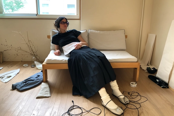 A spontaneous, casually candid iPhone photograph of a young woman reclining comfortably on a low, simple wooden-framed sofa with neutral-toned cushions inside a minimalist Japandi-inspired creative studio space. She wears a thoughtfully deconstructed black cropped tee layered over a loose white top, paired with a gently gathered black wide-legged skirt evoking relaxed silhouettes instead of a ruffled miniskirt. On her feet, she dons off-white tabi-style shoes with soft wool socks offering subtle warmth and texture reminiscent of leg warmers and ballet slippers. Her legs rest naturally, loosely entangled with thin black headphone cables casually draped over warm wood flooring strewn with simple folded fabric scraps and minimal canvas panels leaning against serene warm beige walls. The room is softly illuminated by diffused natural light filtering through large windows, casting gentle shadows that emphasize tactile wood grains, subtle fabric textures, and natural skin details including minimal makeup and gently tousled hair loosely pinned back. The photo’s framing is informal and slightly tilted, enhancing the casual intimacy and spontaneous realism typical of an iPhone candid snapshot, with sparse dried plants and ashtrays discreetly placed to echo the original’s creative disorder in a restrained minimalist environment.
