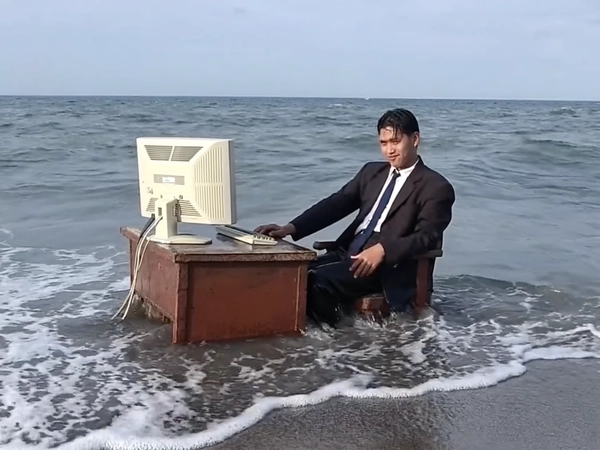 With waves lapping rhythmically against the shore, a young businessman sits calmly waist-deep in the swirling seawater, dark suit dampened by the tide as he rests one hand on a heavy, rust-worn office desk planted directly in the ocean. An old beige CRT monitor perches defiantly atop the warped wooden surface, its cables trailing into the shallow surf below. The photo has a grainy quality, suggesting it may be a still from a video or taken with an older digital camera. His neatly combed hair glistens with droplets, and a faint smirk plays on his lips beneath the muted overcast sky. Quick snapshots create a soft-focus effect at the edges, and muted colors swirl into a nostalgic haze, capturing an absurd twist on remote work culture. A gentle breeze rustles the water around him, the surreal scene blending corporate formality and beach tranquility in deadpan humor—early-2000s digicam snapshot, grainy quality.