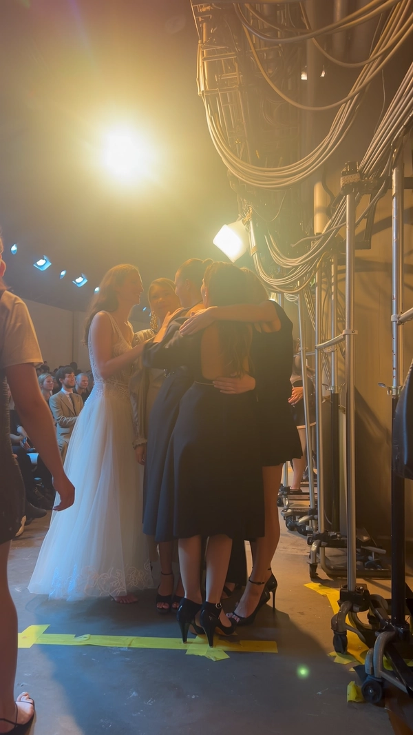 Models cluster just off-runway in a candid embrace, some still poised on glossy heels catching faint motion blur, others barefoot with bare soles touching the gritty, tape-marked concrete floor. Their couture garments spill softly, illuminated by diffused amber haze filtering through industrial fluorescents and scattered tungsten sconces overhead. A low-key soundtrack hums in the background as the camera catches the intimacy of smiles and close-pressed cheeks against a backdrop of coiled cables, rolling racks, and half-lit corridor walls. The iPhone’s sensor renders the soft haze as a gentle bloom around warm bulbs with realistic digital noise in the shadows; fabric textures—from delicate lace motifs to leather straps—remain tactile and nuanced. The composition stretches from an aisle angle just behind the runway’s edge, the frame slightly off-kilter, evoking the transient energy as crowds murmur behind protective barriers and assistants bustle quietly. The informal yet cinematic moment exudes fresh editorial warmth—contemporary runway snapshot, shot on iPhone