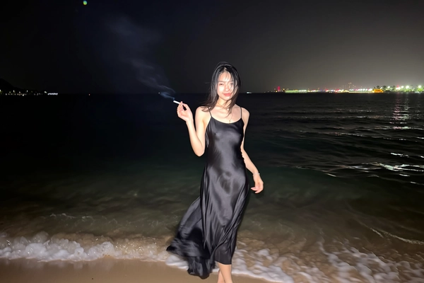 A soft iPhone pop flash fractures the velvet darkness, revealing an elegant woman of East Asian descent standing waist-deep at the shore where gently rippling waves caress her bare feet. Her black satin slip dress flutters lightly in the seaside breeze, shimmering subtly amid faint lens speckles as damp strands of hair frame her serene face. Holding a cigarette with effortless grace, her calm eyes meet the camera's gaze beneath night’s vast expanse, where distant neon city lights ripple faint reflections across the ink-black ocean behind her. The ample negative space cradles this moment, the dress’s blurred hems swirling softly against sharply defined poise—timeless, unembellished, quietly magnetic. —flash-lit elegant beach snapshot, captured on iPhone.