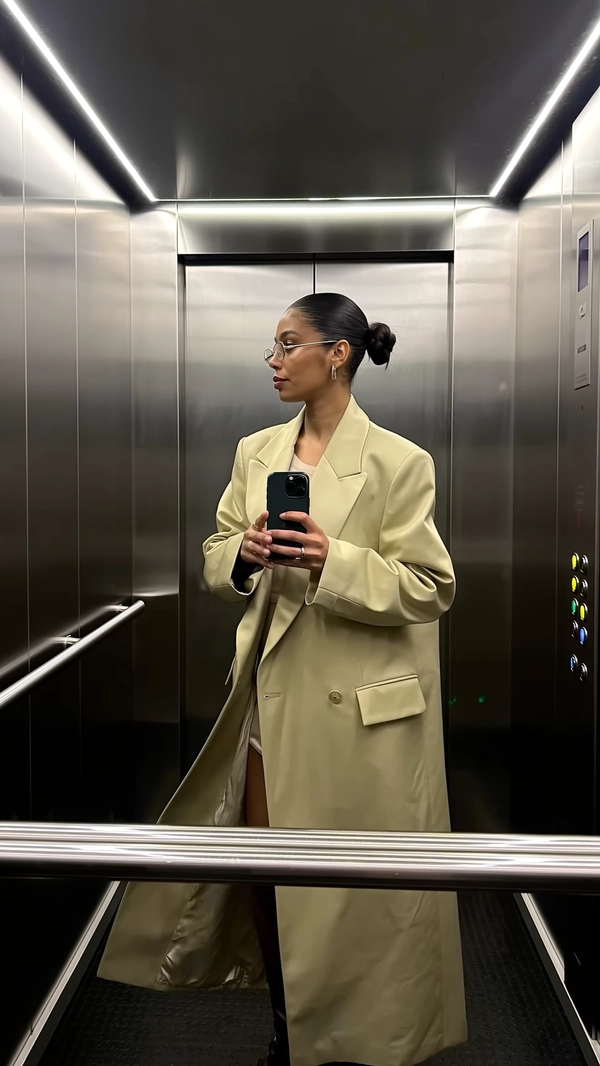 Cool white LEDs slice sharply from a mirrored ceiling, forming a crisp triangle above her head as she stands in a mirror-draped elevator box. Her structured, long beige coat sways subtly around a high slit, the satin sheen catching faintly under the stark glow. Thin rectangular glasses rest deliberately on her nose, framing a smooth, unbothered face whose sleek bun tucks neatly at the nape. With the phone poised low against her waist like a practiced prop, the slight barrel of the lens warps the slim fabric weave of the gabardine coat and the fingerprint smudges on the glass. Her gaze is caught mid-screen, caught between focused and detached, a quiet confidence radiating with every subtle shift as the elevator hums softly, doors inching closed in slow motion—elevator mirror capture, urban chic on iPhone