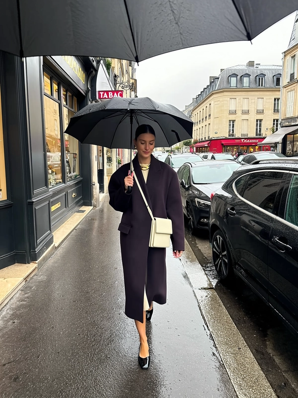 She strides down a narrow rain-slick sidewalk, a black umbrella centered above her calm face, dark plum oversized overcoat with broad shoulders layered over cream silk dress hem peeking delicately below; chunky gold necklace glints subtly while a crisp cream crossbody bag hangs structured at her right hip. Her sleek low bun and pointed black heels add clean geometry to the silhouette, eyes cast softly downward in serene composure. Classic rain-darkened wooden storefronts stretch to her left, wet black cars lined along the opposite side gleam under muted overcast light. In the distance, a pale Haussmann façade pulses with quiet elegance beneath a red ‘TABAC’ sign, misty reflections doubling the scene on the soaked cobblestones. The iPhone’s gentle sensor grain texturizes wet fabric beads and window glass droplets, while the portrait-framed shot carries a slight downward tilt, catching her full stride mid-step and the umbrella rim cutting across the top edge in asymmetrical balance—rainy city snap, shot on iPhone