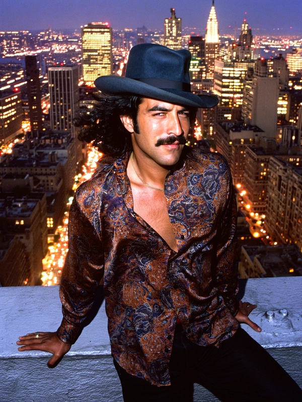 Bathed in the hypnotic glow of sodium-vapour city lights, a Latino man perches on a weathered rooftop ledge, city skyscrapers outlined with flickering amber and cobalt hues sprawling behind him. His shaggy dark hair tumbles beneath a wide-brimmed fedora casting a playful shadow across his forehead, complementing the perfectly groomed mustache that accentuates the teasing curl of his lips. The raised eyebrow hints at confident amusement, the slight squint catching the shimmering neon reflections off his steel-blue eyes.

His paisley shirt, a swirling riot of burnt orange, deep purple, and faded teal, clings softly to his frame, the fabric’s worn cotton texture visible through the subtle grain of old 35 mm stock. The collar is open, revealing a hint of a gold chain absentmindedly catching the city’s dim light. The breeze teases the shirt’s fabric and ruffles raven hair in motion, frozen mid-flutter in a deliberate, languid pose that balances relaxed cool and magnetic intensity.

The shot, composed with a slightly grainy 50 mm lens at eye level, frames him against the sprawling urban maze below. The warm sodium vapour creates pools of saturated amber contrasts with the cooler indigo shadows that shape his silhouette and the rooftop’s gritty concrete. Highlights catch on skin pores and the soft sheen of the shirt’s fibers, merging tangible reality with nostalgic moodiness.

Captured in that exact moment between night’s pulse and contemplative solitude, the image channels the cinematic style of the era’s New York street portraits, echoing an unspoken narrative of urban charisma and quiet rebellion. The analog film grain weaves authenticity through the timeless atmosphere.

—late-70s / early-80s cinematic photograph, authentic film grain.