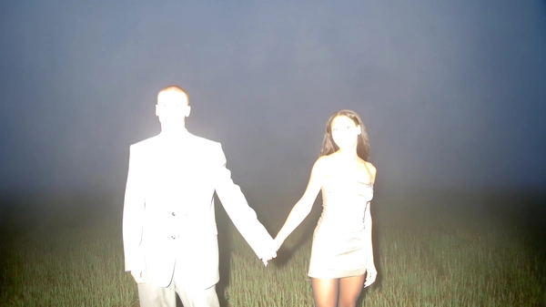 A candid dawn snapshot in a misty field, captured with an intense iPhone flash causing complete bleaching of two mixed-race figures — one wearing an oversized suit, the other in a sheer dress with tights. Their faces and clothing details are obscured by harsh overexposure, leaving only faint, ghostly outlines. Their intertwined hands and confident stances remain discernible, highlighted against the soft, darkened misty background. The spontaneous, slightly imperfect framing typical of an amateur flash photo emphasizes a surreal, ethereal atmosphere.