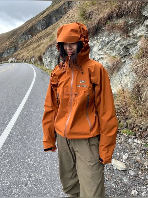 A candid iPhone photography style image capturing a beautiful Asian woman standing relaxed beside a mountain road, wearing a rust orange windbreaker jacket with an Arc'teryx logo, paired with loose khaki pants, embodying the practical and rugged gorpcore aesthetic. Her posture is casual with arms loosely at her sides, her face softly obscured by the jacket hood pulled up casually, showcasing neutral expression. The scene is set along a rugged mountainous roadside lined with textured rocky formations and dry wild grass, under slightly diffused daylight with partial cloud cover typical of spontaneously captured mountain ambiences. The jacket fabric shows authentic creases and subtle dirt marks, natural folds catching the soft light. Surrounding terrain textures of rocks and wild grasses appear richly detailed with visible imperfections. The framing is slightly tilted, enhancing the genuine, intimate feel of a spontaneous moment. The overall style aligns closely with a trendy technical outdoor aesthetic, emphasizing authenticity and casual mountain realism.