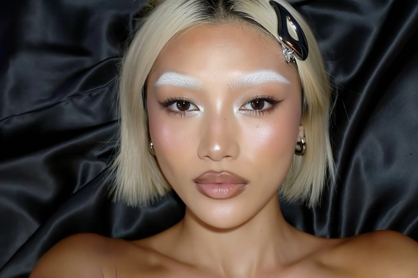 An authentic, close-up overhead iPhone selfie perspective of a young woman with perfectly symmetrical features, naturally bleached eyebrows blending subtly into her skin creating a pale blonde effect. Her makeup showcases glittering lashes and luminous glass-skin glow with muted, glossy nude lips and minimal eye accents. She lies on luxurious black silk fabric, wearing a sleek chrome hairclip that adds a contemporary, understated metallic accent. Her expression is calm and neutral, directly engaging the camera with soft, natural indoor lighting highlighting realistic skin texture, makeup sheen, and subtle imperfections. The background is a simple black silk surface, enhancing the sophisticated yet casual aesthetic typical of authentic handheld iPhone selfies.