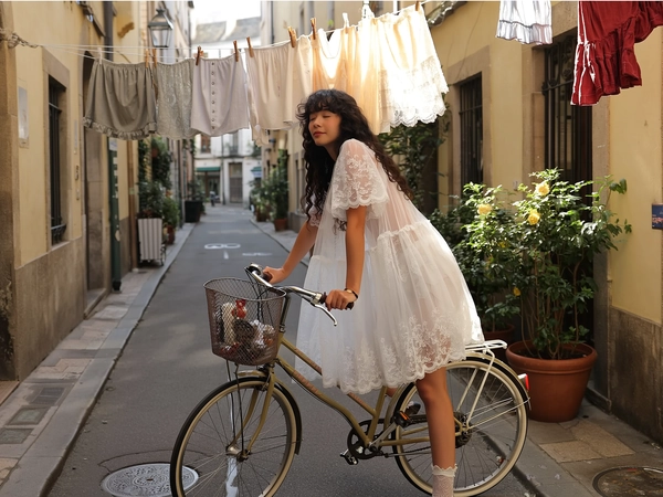 A softly whimsical, intimate moment casually caught on an iPhone, portraying a young woman with naturally curly black hair styled loosely, dressed in a sheer lace babydoll dress characteristic of Jenny Fax, delicately layered with subtle scalloped edges and modest white lace-trimmed socks. She leans gracefully on a vintage bicycle along a quiet city street lined with charming potted plants. Sunlight filters warmly and softly through gently swaying hanging laundry above, illuminating the intricate lace transparency, softly textured fabric drape, and the aged patina of the street ambiance. The scene is framed from a slightly elevated and gently tilted angle, capturing her peaceful expression and the tender blend of playful romanticism with refined vintage charm, all woven seamlessly in a pastel-hued, quietly feminine aesthetic.