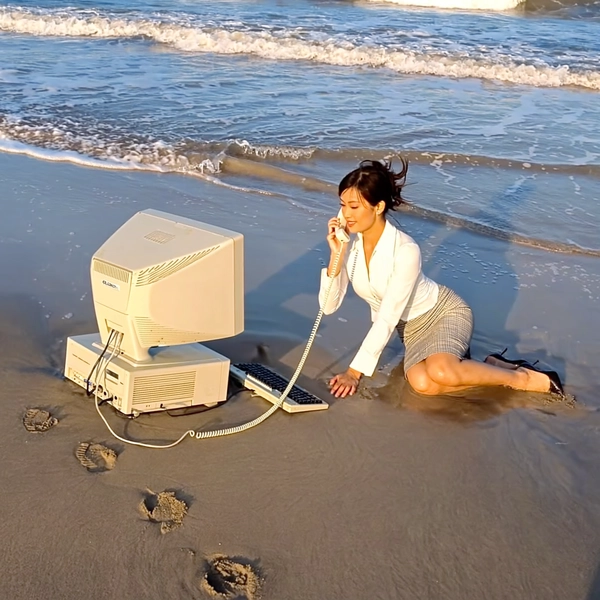 With waves lapping rhythmically against the shore, a woman kneels gracefully in the warm sand, clad in a fitted white blouse and checkered pencil skirt, calmly holding a corded landline phone to her ear as she faces a beige CRT monitor and chunky keyboard balanced on the wet sand. The computer tower tilts slightly beside her, the white telephone wire snaking elegantly across footprints imprinted in the sand. The photo has a grainy quality, suggesting it may be a still from a video or taken with an older digital camera. Her neatly tied hair flutters gently in the soft morning breeze, her serene expression untouched by the absurd twist on remote work culture juxtaposed against the beach tranquility. Quick snapshots create a soft-focus effect at the edges, and muted colors swirl into a nostalgic haze, capturing the unlikely marriage of corporate formality and coastal peace. Sunlight casts golden shadows that bring out every texture, from fabric weave to fine grains, while the surf lazily swirls nearby—the early-2000s digicam snapshot, grainy quality.