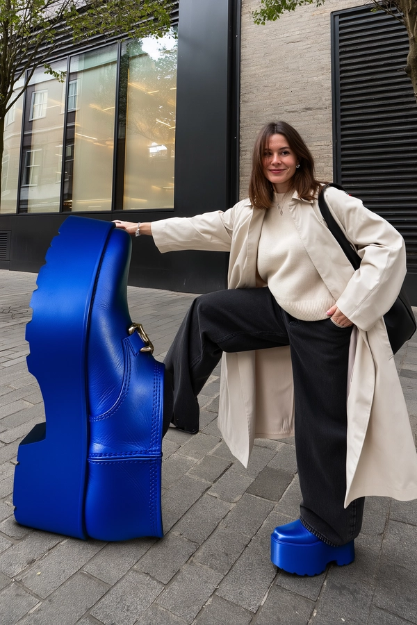 A spontaneous, candid iPhone-style photo of a petite woman climbing a giant cobalt blue platform boot positioned on a picturesque city sidewalk, styled as an urban sculptural landmark. She wears the exact same cobalt platform boot in standard size on her right foot, visible with detailed leather texture, subtle creases, and glossy finish. Her fashionably casual outfit features a light beige trench coat over a soft knit cream sweater, slim black jeans with natural folds, and minimal silver jewelry. The boutique backdrop displays large glass windows framed by sleek matte black metal, subtly reflecting soft daylight. Natural outdoor daylight filters gently through nearby trees, casting soft, nuanced shadows on the textured stone pavement and boutique facade. The image captures skin texture with visible pores and fine hair strands, detailed fabric weaves on clothing, and realistic leather grain on the boots. The photo is informally composed with a slight tilt, the boot and woman off-center, evoking a candid, spontaneous iPhone snapshot blending surreal scale with natural urban elegance.