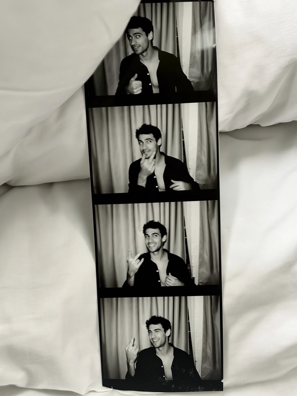 A vertical black-and-white photostrip lies softly atop white bedding, fabric folds gently framing its worn surface. Two men appear throughout the four frames: the first shows them both gazing thoughtfully off-camera, their unbuttoned shirts lending casual ease. In the second, one man winks playfully, sparking spontaneous connection. The third captures matching hand gestures, their subtle movements synchronized yet relaxed. The final frame reveals a faint smirk crossing their lips, completing a story of nuanced expressions. Behind them, neutral beige curtains ripple subtly, grounding the setting in classic photobooth authenticity. The strip carries gentle sensor grain, faint fingerprints, and slight creases, all enhanced by muted contrast and soft iPhone flash reflections. Slightly skewed and casually arranged, the moment feels intimately lived and effortlessly timeless—contemporary photobooth snapshot, shot on iPhone.