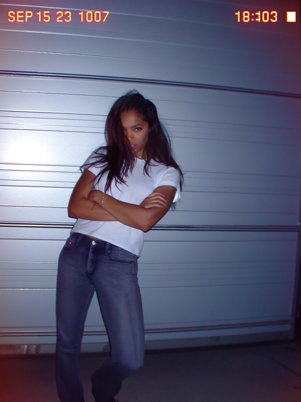 A sudden pop from the on-camera flash throws hard shadows against the plain metal garage door, catching her tucked-in white tee and tailored jeans in a soft but harsh tungsten glow. Her arm crosses protectively as if waiting, the edges around her blurred just enough by subtle camera shake and pixel fringing. The faded tungsten light swirls with muted pastel noise across the rough metal skin, while a quick tilt skews the composition like a rushed snap. SEP 15 2003  18:47 blinks in the corner, alongside a faint battery icon. JPEG blocks crunch unevenly near the frame's edge, and the photo quality is low-resolution and somewhat grainy.—early-2000s digicam snapshot, grainy low-res quality