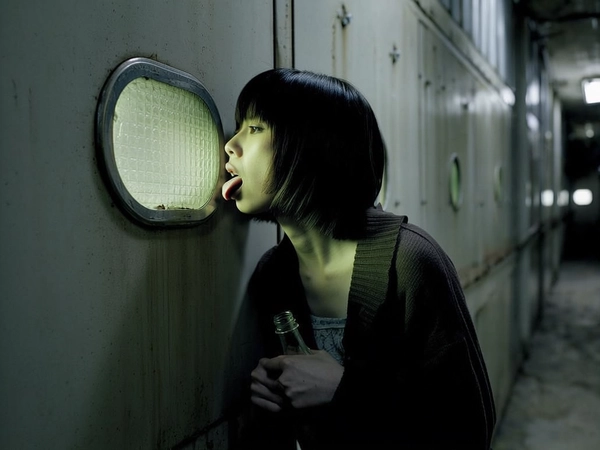 In the dim, claustrophobic corner of a decaying industrial corridor, a young Japanese woman leans toward a grimy metallic wall, her short, blunt bob haircut framing a face taut with desperate curiosity. Her tongue presses hauntingly against a small fogged glass window embedded in the metal, eyes intense beneath low, greenish-brown light that seeps into rust-stained surfaces. She clutches a cloudy, half-empty glass bottle, her trembling fingers frozen mid-gesture, encapsulating a raw moment suspended between hope and fear. The surrounding atmosphere hums with grainy texture—softly blurred institutional walls fading into shadow, evoking the unsettling stillness of a late 90s psychological thriller. Muted, desaturated hues blend with directional light, pushing edges into an eerie dreamscape that feels both intimate and disturbingly cold. The tactile coldness of metal contrasts with the fragile warmth of skin, captured through the vintage Leica lens softness, inviting the viewer to dwell within this fragile, anxious gaze.