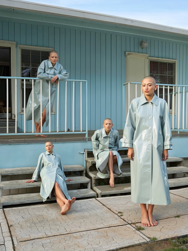 A surreal, spontaneously candid iPhone-style photograph featuring a woman with a shaved head and delicate pearl-studded eyebrows, duplicated multiple times throughout a pale blue motel courtyard. Each version wears the exact same floor-length glossy vinyl trench coat, standing, leaning casually over metal railings, sitting barefoot on worn concrete steps, and walking barefoot across cracked tiles, creating a serene geometric pattern. The natural daylight softly illuminates her detailed skin texture and the shiny, flexible vinyl fabric, casting gentle shadows that enhance depth and realism. The composition employs varied scales and perspectives—some close, some distant—showcasing relaxed, unposed expressions and movements that feel effortlessly authentic, accentuating the surreal repetition while retaining the spontaneity and texture-rich aesthetics typical of an informal iPhone snapshot.
