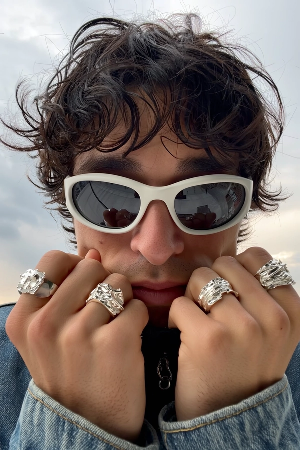 He tucks his chin behind a collar pulled tight, short dark hair tousled from a lazy day, the white curve of his sunglasses catching a crisp city skyline like a fractured mirror. Stacked silver rings with molten, sculptural shapes glint along his fingers, cool and fluid as they press denim-knit fabric closer to the skin, where pores and a faint smudge clutter the lens edge. An overcast afternoon filters in, lending a muted glow that softens the geometry of his features. The frame dips in a casual Dutch tilt, center-focused on rings that catch stray flare while the reflection holds secrets beyond the glass— close-up captured on Iphone, hand-face jewelry focus