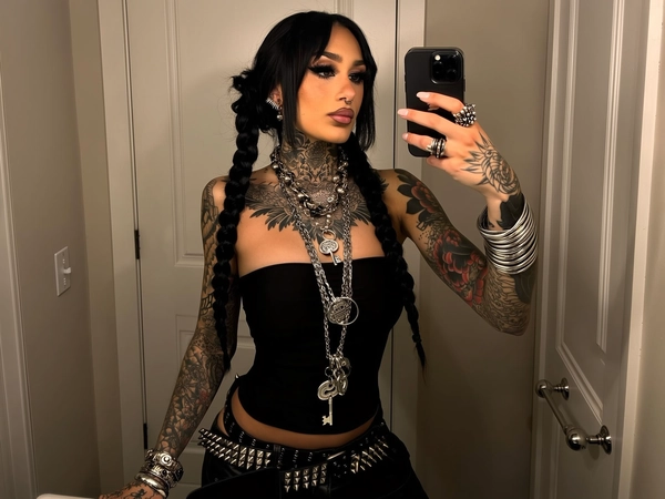 A young woman with jet-black hair styled in two long, messy braids stands taking a mirror selfie in a dimly lit room with white paneled doors. Her skin shows rich detail and texture, fully tattooed across her arms, chest, and neck with intricate floral and ornamental designs. She wears a tight black strapless top and black low-rise pants accented with layered studded belts. Heavy silver jewelry — thick chain necklaces with large key pendants, stacked spiked bracelets, and chunky rings — adorn her. Her makeup is bold: thick black eyeliner, long lashes, and glossy lips. Facial piercings include a septum ring and multiple studs. Warm bathroom lighting softly highlights subtle shadows and natural skin imperfections, giving an authentic, spontaneous iPhone mirror selfie feel.