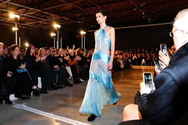 A commanding female model glides down the runway wearing a deconstructed evening gown rendered in sheer, layered chiffon and structured satin panels that catch the light with subtle translucence. Her sleek, wet-effect hair slicks close to her scalp, contrasting with minimal, natural makeup that highlights her sharp cheekbones and focused expression. The fashion royalty seated in the front row watch intently from plush benches set beneath an industrial loft ceiling with exposed beams and visible rigging grids. Overhead, a spectrum of amber and cool white spotlights splash warmth against the translucent fabrics, creating soft light blooms and pools of shadow on the concrete runway marked with taped sightlines. The iPhone capture registers slight digital noise in the darker corners and a gentle motion blur on the gown’s flowing train, lending a candid immediacy to the moment. Shot from a front-row oblique angle just as the model advances past attentive spectators wielding phones and whispered applause, the composition teeters naturally with a subtle hand-held tilt—contemporary runway snapshot, shot on iPhone