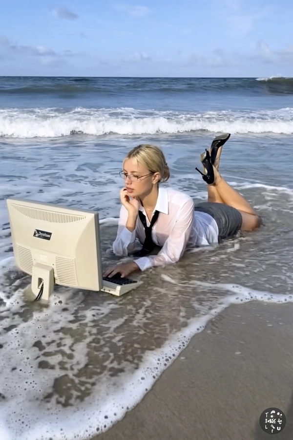 With waves lapping rhythmically against the shallow surf, a blonde woman lies on her stomach at the ocean's edge, her soaked white office shirt clinging beneath a loosely knotted tie and grey pencil skirt, the black high heels dangling from her raised, bare feet. The midday sun casts a bright, cloudless light that reflects softly off the clear water swirling around her arms and the beige CRT monitor and full-sized keyboard half-submerged in the surf. The photo has a grainy quality, suggesting it may be a still from a video or taken with an older digital camera. Her neatly tied-back hair glistens with wetness, rimless glasses perched precisely, her thoughtful gaze fixed on the screen amidst this surreal twist on remote work culture. Quick snapshots create a soft-focus effect at the edges, and muted colors swirl into a nostalgic haze, the gentle ocean breeze teasing the wet fabric and rippling the still water, merging the rigidity of office life with beach tranquility—early-2000s digicam snapshot, grainy quality.