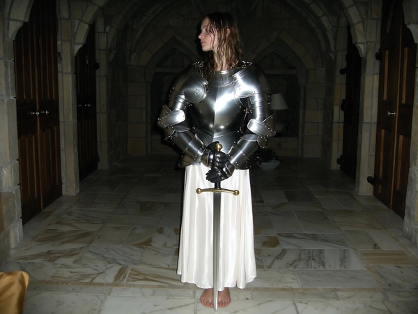 A wide, full-body, eye-level shot from an early 2000s-style digital camera explicitly capturing a young woman standing barefoot on a marble floor inside a darkened room evocative of a medieval castle chamber at night. She wears polished full medieval armor elegantly layered above a silky nightgown, with damp hair loosely falling over her chestplate. She holds a sword vertically in front of her with both hands, head turned thoughtfully sideways, gaze directed away from the camera, reflecting calm knightly composure. The harsh direct flash of the vintage digital camera highlights the shining metal armor textures and soft fabric details amid the muted, darkened indoor environment with heavy shadows. The overall fragile and solemn atmosphere conveys a surreal medieval-core ambiance blended with contemporary domestic elements. The wide full-body framing at eye-level preserves the knightly elegance and quiet dignity characteristic of early 2000s digital photography.
