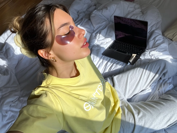 A stylish young woman sits casually on an unmade bed bathed in soft daylight, wearing a pastel yellow oversized T-shirt with subtle white text and cozy light gray sweatpants. Her skin glows fresh beneath glossy deep lavender hydrogel under-eye patches, while her hair is tied back loosely with a scrunchie, complemented by delicate gold hoop earrings. Nearby, a tube of hand cream and an open laptop rest casually atop soft, slightly rumpled sheets. The natural window light gently illuminates her radiant skin and the subtle sheen of the hydrogel patches, enhancing the cozy textures of her loungewear and bedding. Shot from a top-down selfie angle, the framing captures her face, shoulders, and upper torso with realistic iPhone grain, conveying an authentic, relaxed self-care morning moment in a softly lit bedroom scene—skincare selfie, shot on iPhone.