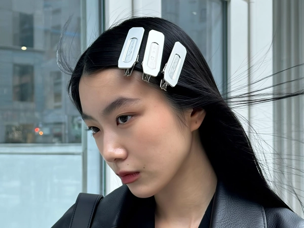 Pristine white rectangular clips line up perfectly above the ears, their sharp edges catching the icy café window glow as a young woman with jet-black straight hair offers a calm, focused stare. The clean contrast between the matte clips and sleek strands frames her clear glass-skin glow, while stray fly-aways soften the calculated feel. A soft leather lapel grazes her neck, minute grain evident, and the shot tilts subtly left, hinting at a high-angle stretch; city pavement blocks faintly blur behind, grounding the cool minimalism in everyday life—authentic iPhone close-up, statement hair-clip focus