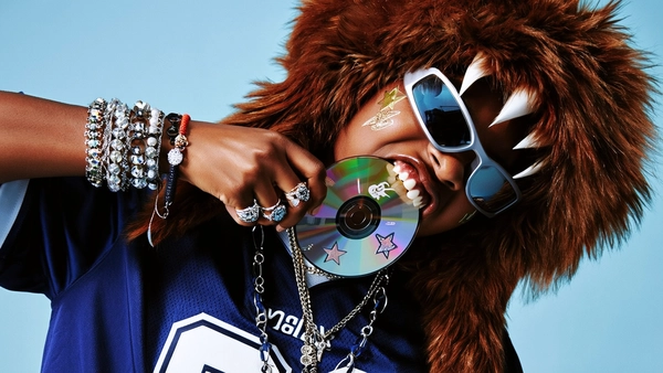 A close-up portrait captures a young Black model wearing a plush brown monster hat, its cartoonish teeth protruding fiercely as they precariously bite down on a reflective CD. Their futuristic wraparound sunglasses shimmer with metallic silver glints, cool and unreadable, reflecting fractured pastel brights that dance across a soft blue backdrop. The Dallas Cowboys jersey anchors the composition with deep navy hues, layered beneath an array of silver chains, beaded chokers, and chunky bracelets that clatter with subtle movement. Star-shaped stickers scatter their cheeks, playful bursts of color carved into the skin with rebellious vitality.

The lighting sculpts their features with a cinematic hardness—key light strikes sharply from above, carving out confident shadows that deepen the Y2K mood, while a soft fill gently caresses the polished textures of metal and fabric. Warm earth tones meet electric pastels in this tangled tableau, rims of light outlining every ring and bracelet as if pulsing to early internet static.

Detail becomes devotion: the woven jersey fibers catch every thread, the CD's mirror slide gleams with fingerprint smudges and light scratches, and skin pores hold the tension of the moment. The tilted frame slices diagonally, compressing energy into a tight, kinetic heartbeat; shot with a 90mm lens at mid-aperture, focus sharp, background gently softened.

Processed with a digital profile echoing trademark high-contrast Y2K editorial—saturated colors break against rich shadows, textures heightened but never slick—an homage to internet-era bravado crystallized in tactile clarity. The rebellious spirit of an age where tech and kitsch collide pulses vividly here.

—high-fashion editorial, hyper-real texture fidelity