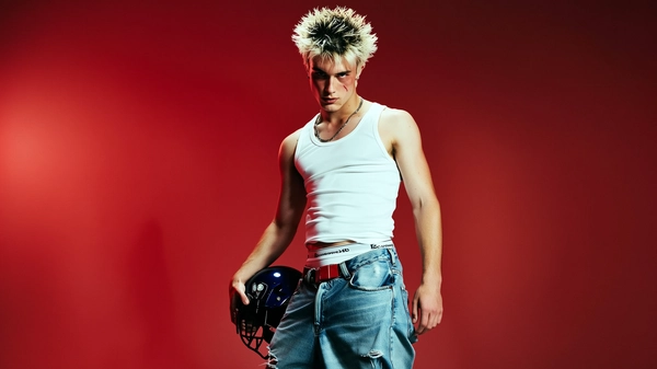 He's framed in a medium close-up beneath a stark red studio backdrop lit with harsh directional lighting that sculpts stylized shadows across the scene. His bleach-blonde hair is spiked with visible dark roots, tousled and rebellious. He wears a crisp white tank top snugly tucked into oversized, heavily distressed denim jeans that sag just enough to reveal the waistband of his boxer briefs, detailed with a bold logo. One hand grips a glossy black football helmet, held low and casual but purposeful. His face tells a story—slight bruising creeps beneath a sharp jawline and past intense eyes locked deadpan at the camera, embodying a rebellious 2000s teen drama vibe. The overall feel is gritty yet styled, raw but controlled—a perfect capture of early-2000s youth culture posing for a fashion editorial that fuses street cred with high attitude. —casual candid early-2000s Y2K snapshot, grainy softness