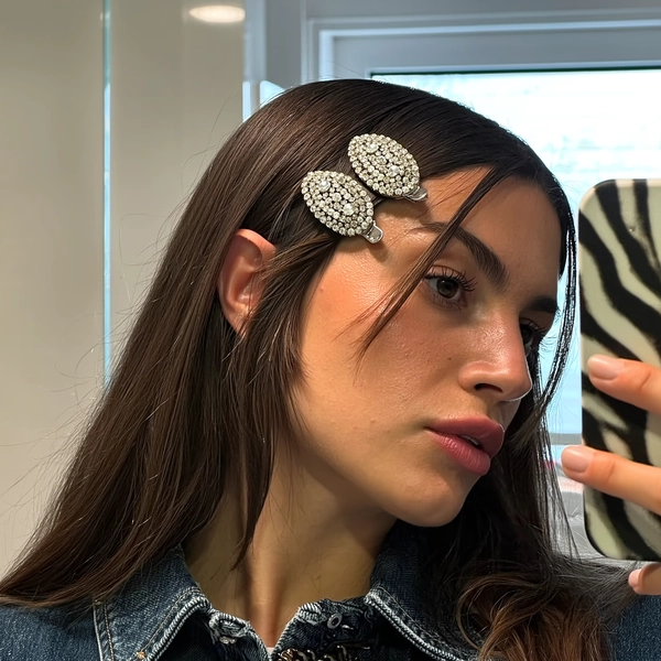 Pearl-encrusted snap clips gleam softly amid sleek chocolate brown strands, framing the centre part with delicate shimmer as they catch the gentle glow from a bathroom window. Her calm, glass-skin face reflects a quiet confidence, lips parted slightly while holding a zebra-print phone case caught in the mirror’s edge. Fine flyaways brush the edges of her dewy cheeks, skin pores evident in the soft light, while her relaxed denim collar peeks in below. The shot tilts just so, a casual mirror selfie that feels both spontaneous and polished, the clips truly stealing the scene. —authentic iPhone close-up, statement hair-clip focus