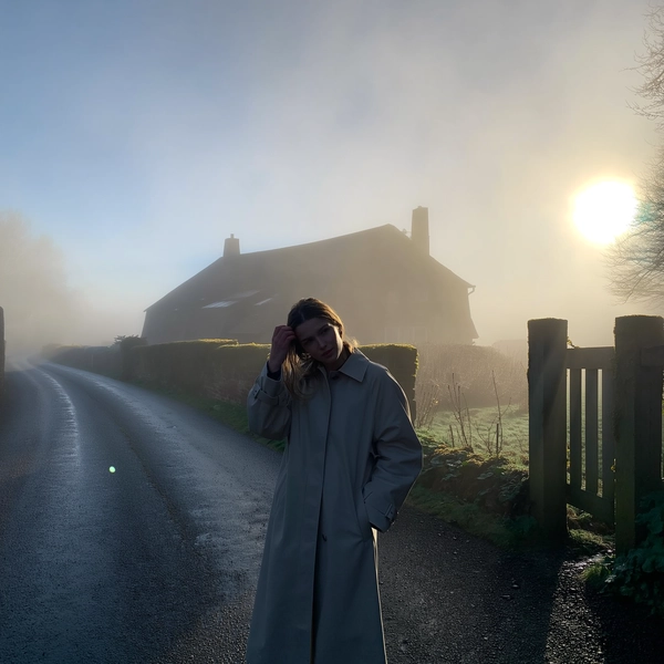 A young woman clad in a sleek, minimalist trench coat stands still on a slick, rain-dark asphalt path winding past a rustic cottage whose outlines blur softly into thick, lingering fog. Her loose hair clings damply to her neck as she tilts her chin slightly forward, breath visible in the cold, and one hand slides into her coat pocket while the other brushes a stray strand from her face. The dense mist swallows color into pale greys and muted greens of moss clinging to gateposts, while a faint, diffused dawn light softly pools through the haze, casting gentle luminance with a subtle lens flare near the blurred sun. The iPhone's grain texture whispers through shadowed corners; droplets bead quietly on wilting leaves, and the faint tap of distant dripping condensation layers the stillness. The shot, caught from chest height with a hand-held, slightly tilted frame, leaves generous negative space above and around her, dissolving the world into quiet solitude—mistbound reverie, shot on iPhone