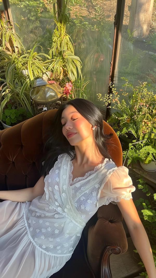 A softly intimate moment delicately revealed in warm, naturally diffused sunlight inside a sunroom lush with gently overgrown greenery, where a young woman reclines gracefully on a deep velvet chaise. She wears an ethereal Jenny Fax white sheer lace dress, intricately detailed with scalloped edges and delicate floral appliqués, featuring softly puffed sleeves and subtle ruffled layering. Her flowing black hair cascades loosely around her shoulders, complementing minimal accessories and fresh, natural makeup that highlights her serene, introspective expression. Golden shafts of afternoon light illuminate her face, enhancing the translucent lace textures and the rich velvet fabric beneath her. The gently weathered, plant-filled space adds quiet vintage charm, while the composition is framed from a subtle elevated angle with a slight tilt, creating a harmonious balance of tender femininity and effortless whimsical intimacy.