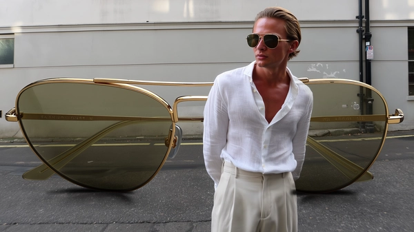A spontaneous, candid iPhone-style photograph of a male-presenting model with strong facial symmetry and olive-toned skin, his brushed-back blond hair softly textured with individual strands visible. He wears a fitted white linen shirt with a delicate natural weave and muted cream trousers, standing relaxed with a slight recline across the frame. Over his eyes sit a true-to-scale pair of classic golden aviator sunglasses, their reflective lenses capturing soft urban reflections. Beneath him extends a dramatically oversized pair of aviator sunglasses, meticulously detailed with gleaming golden metal frames showing slight surface imperfections, fine screw joints, and glass lenses tinted subtly to mimic the smaller pair. The enormous glasses rest naturally across a sunlit, lightly weathered concrete urban sidewalk featuring gentle cracks and faint paint markings. The ambient outdoor daylight is diffuse and gentle, casting realistic soft shadows that play subtly on the model's textured skin, the fine fabric of his outfit, and the reflective frame surfaces of both sunglasses. The composition is casually informal, slightly off-center with a mild tilt typical of spontaneous iPhone snapshots, seamlessly integrating the surreal oversized accessory to create an authentic yet imaginative visual contrast within a quiet urban setting.