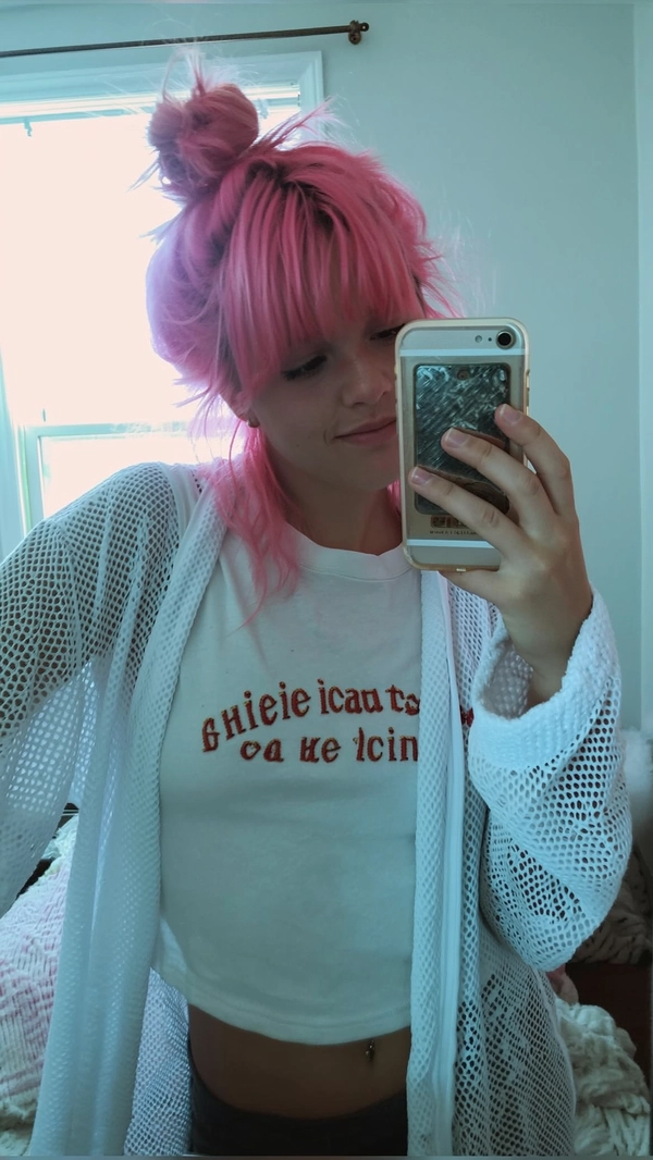 A casual, spontaneous selfie of a young woman with vibrant pastel pink hair styled into a messy bun, wearing a white mesh robe loosely over a crop top featuring ironic text, standing in front of her bedroom mirror. The room is softly illuminated by sun-bleached daylight streaming through the window, creating a washed-out, pale-toned atmosphere characteristic of 2016 Tumblr selfies. Muted color saturation and softened contrast dominate the image, with cool-toned shadow hues subtly mixed with a muted warm overlay, maintaining authentic mid-2010s aesthetic textures. Visible textures include the delicate mesh fabric, slightly tousled loose hair strands, detailed skin textures with faint imperfections, and casual relaxed expression. The framing is informal and off-center with a slight tilt, evoking the genuine spontaneity and candid intimacy of mid-2010s smartphone photography.
