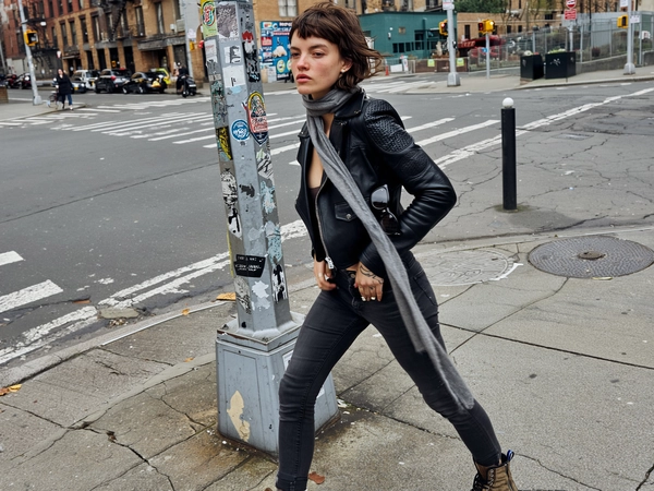 A tall, slim young woman strides across a cracked, dirty sidewalk at a gritty New York street corner, captured in a spontaneous mid-close iPhone shot with a slight off-center tilt. She wears a fitted black leather jacket with cracked texture, left open over worn black skinny jeans and scuffed combat boots, all deeply textured and lived-in. A long, thin gray scarf hangs loosely around her neck, fluttering lightly in a soft breeze. Her short, choppy brown hair is naturally unstyled, uneven with subtle flyaways, framing her face with an androgynous edge. Beside her, a weathered lamppost covered in peeling stickers and chipped paint blends into urban decay.

Her tired, detached expression faces away from the camera, eyes unfocused, conveying muted emotion. Skin texture is raw and authentic, showing visible pores, slight redness, and natural shadows under the soft, overcast daylight that gently sculpts her jawline and cheekbones. Both hands display delicate, minimal linework tattoos, fingers loosely curled as she moves. The composition feels immediate and unpolished, with a faded, desaturated color palette and gritty texture echoing a Tumblr-era indie sleaze vibe, resembling Alexa Chung's effortless messy chic. A large black sunglasses accessory is folded and tucked into her jacket pocket, adding subtle edge to the look.