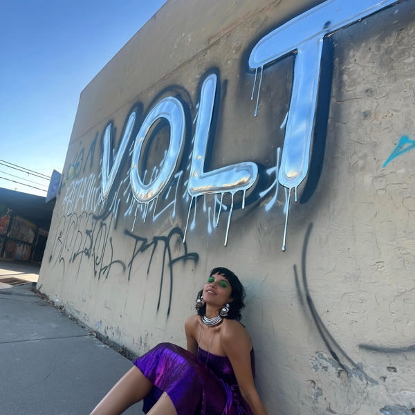 A candid urban portrait of a woman sitting beneath a massive chrome-letter graffiti mural reading:
"VOLT"
She has striking green eyebrows and wears a deep purple wrap skirt, her reflective silver jewelry catching subtle silvery highlights from the metallic graffiti. The scene is set against a weathered concrete wall with faint graffiti textures around the chrome letters. Natural daylight softly illuminates the scene, enhancing the reflective surfaces and casting gentle shadows. The photo is taken with a casual, slightly tilted angle from eye level, typical of spontaneous iPhone photography, capturing authentic street ambiance and raw urban textures with realistic imperfections.