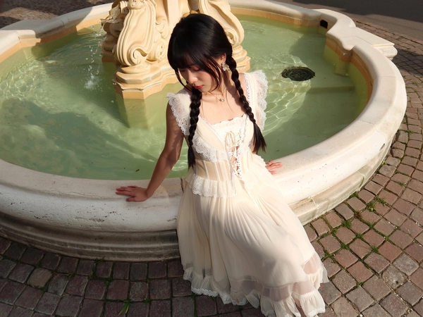 A softly intimate moment captured with the natural ease of an iPhone, featuring a young woman seated gracefully on the edge of an old town square fountain. She wears a whimsically feminine Jenny Fax-inspired cream lace corset top with intricate scalloped lace trims and delicate embroidery, paired with a flowing, sheer layered skirt that drapes gently around her. Her long black braids cascade smoothly over her shoulders, complementing the serene, introspective expression and subtle, fresh makeup. Soft shadows play across the textured stone fountain and worn cobblestones, bathed in warm, diffused afternoon light that caresses the translucent fabric and lace details. The composition is framed from a gentle elevated angle with a subtle tilt, balancing generous negative space to evoke a tranquil vintage romance infused with delicate playfulness.