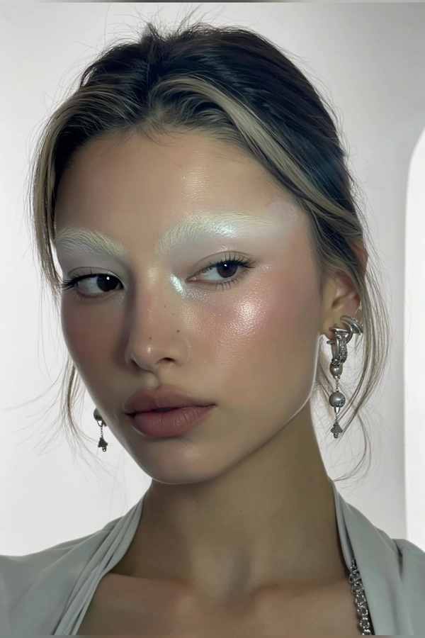 A close-up, authentic iPhone selfie-style portrait of a young woman with naturally bleached eyebrows softly blending into her skin, showcasing sculpted cheeks with subtle shimmering highlight that adds dimension. Her makeup remains natural with muted matte lips and minimal eye makeup emphasizing faint icy or silver tones. The lighting is sterile and soft, casting gentle, diffused shadows that highlight the shimmer on her skin while keeping the overall mood calm. She gazes neutrally but engagingly into the camera, her hair naturally framing her face. The background is a smooth, seamless white environment without visible corners, evoking a minimalist, modern aesthetic akin to a softly lit white cube. She wears a simple, light-colored top with understated silver accessories, reinforcing the effortless and contemporary vibe of the iPhone selfie.