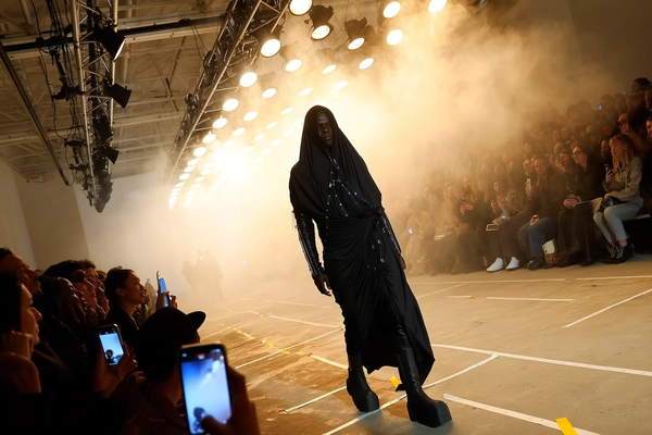 A figure cloaked head-to-toe in Rick Owens’ signature dystopian black drapery emerges from a dense veil of theatrical fog, the angular, layered silhouette cutting sharply against the pale mist. The model’s leather harness catches sparse glints from overhead tungsten bulbs, contrasting with matte oversized boots grinding the runway’s gritty concrete streaked with taped sight-lines. Audience faces, mostly shadowed but punctuated by the soft blue glow of raised smartphone screens, press forward in a chaotic crush along narrow benches in an industrial loft space evoking brooding minimalism. A grid of amber PAR cans above creates a warm, diffused glow that gently swells around the fog, the iPhone sensor rendering a distinct bloom that blurs edges while capturing the raw fabric textures and the model’s steely expression. The shot is taken from an oblique front-row angle with a slight tilt, lending a sense of immediacy and unsettled anticipation as the runway’s stainless steel rigging looms overhead and a phone flash ignites briefly behind the crowd. —contemporary runway snapshot, shot on iPhone
